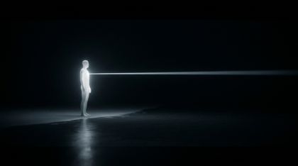 A lone luminous figure stands at the edge of an infinite black ocean and, from its heart, casts a single steady beam into the dark — not a searchlight seeking anything, but a quiet “I am here,” offered without demand.
